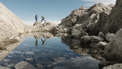 Zwei Wanderer springen zwischen Felsen über einen Bergsee
