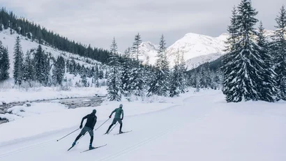 Zwei Langläufer im Winterwald mit schneebedeckten Bergen im Hintergrund