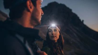 Frau mit Stirnlampe schaut bei Dämmerung Mann beim Wandern vor Berg an