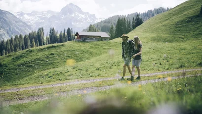 Paar wandert auf einem Bergweg mit Hütte und schneebedeckten Bergen im Hintergrund