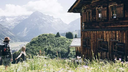Zwei Wanderer vor einer Holzhütte in den Bergen mit Bergwiese im Vordergrund