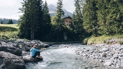Frau sitzt am Flussufer in den Bergen nahe einem Holzhaus