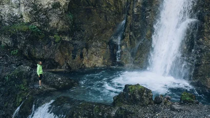 Person in grüner Jacke steht an felsigem Wasserfall mit blauem Wasser