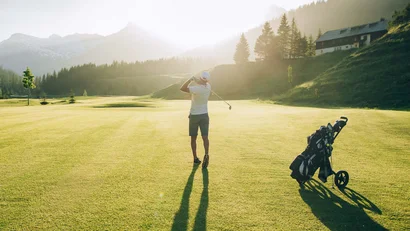 Golfer schlägt Ball auf sonnigem Platz mit Bergen und Haus im Hintergrund