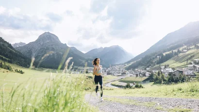 Frau joggt auf einem Weg im grünen Tal mit Bergen und Dorf im Hintergrund
