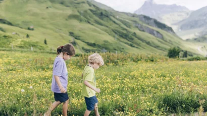 Zwei Kinder laufen barfuß durch eine blühende Wiese in den Bergen