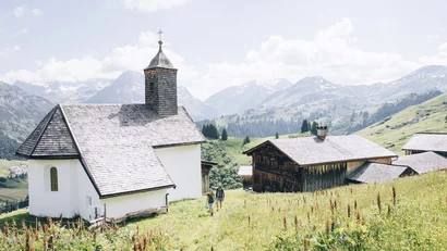 Kleine Kirche und Holzhaus auf Wiese mit Bergkulisse und zwei wandernden Personen