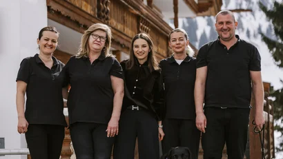 Five people in black clothes with two dogs in front of a wooden house in the snow